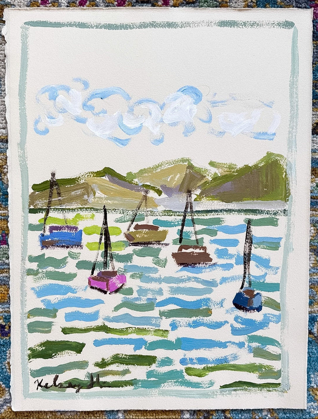 A small rectangular painting or print showing a harbor scene with sailboats, blue water, green hills, and white clouds, placed on top of a patterned blue and gold rug.
