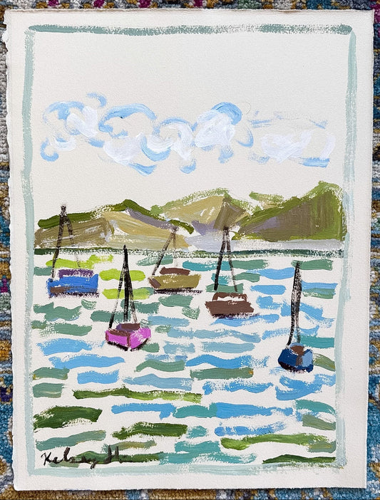 A small rectangular painting or print showing a harbor scene with sailboats, blue water, green hills, and white clouds, placed on top of a patterned blue and gold rug.
